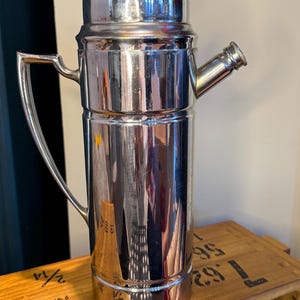 May include: A vintage chrome cocktail shaker with a red cap and a spout. The shaker has a handle and a lid. The chrome finish reflects the surrounding environment. The shaker is cylindrical and sits on a wooden surface. A classic barware item.