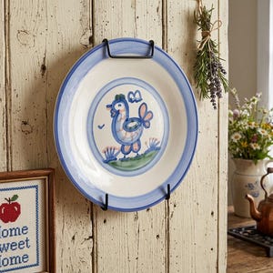 May include: A round ceramic plate with a blue rim and a hand-painted blue and peach rooster illustration in the center. The plate is held by a black metal wall mount against a light-colored wooden plank wall, featuring a central bird motif.