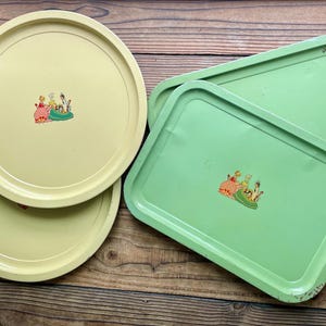May include: A collection of vintage metal trays in shades of yellow and green. The round trays are light yellow, while the rectangular trays are a pastel green. Each tray features a small, colorful illustration of figures in a garden setting.