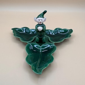 May include: A glossy green ceramic serving dish shaped like three joined holly leaves. A small, smiling elf figure with a pointed green hat and white gloves stands in the center of the dish, holding its hands together in front of its chest.