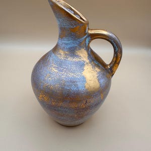 May include: A ceramic pitcher featuring a rounded body and a curved handle. The surface is finished with a mottled texture, displaying a blend of metallic gold and dusty blue tones that create a layered, weathered appearance across the entire exterior.
