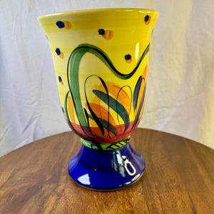 May include: A ceramic goblet with a flared rim and a pedestal base. The upper portion is yellow with hand-painted green swirling lines, orange petal shapes, and small blue dots. The base is a solid, glossy cobalt blue with a green accent ring.