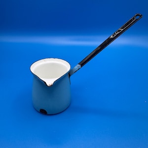 May include: A white enamel coffee pot with a long, slender black handle. The pot features a cylindrical body with a small pouring spout and a black rim. The interior is white, and there is a small dark mark on the lower side of the exterior.