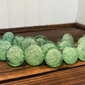 May include: A collection of round, light green glass knobs featuring an internal bubble pattern. Each knob has a smooth, spherical surface with visible air pockets trapped within the translucent glass, creating a textured appearance throughout.