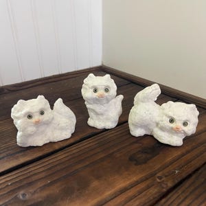 May include: Three white cat figurines with textured surfaces. Each cat has green eyes and a pink nose. One cat is sitting, one is standing, and one is in a playful pose. The figurines are displayed on a dark wooden surface.