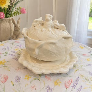 May include: A cream-colored ceramic tureen with a matching saucer, featuring a lid adorned with three sculpted rabbits. The bowl and saucer have a textured, leaf-like design, and the entire set has a glossy finish, presenting a cohesive look.