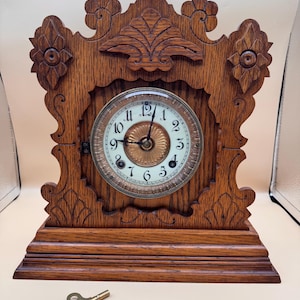 May include: A brown wooden mantel clock with an ornate carved frame and a circular white face featuring black Arabic numerals. The clock has a central gold-toned decorative dial and two black hands. A small brass winding key is positioned below.