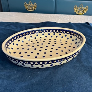 May include: Oval ceramic serving dish with a cream-colored interior and a blue dotted pattern. The rim features a scalloped design with blue dots. The dish is set against a dark blue background.
