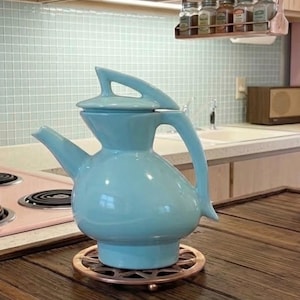 May include: A light blue ceramic teapot with a rounded body, a tapered spout, and a matching lid featuring a triangular handle. The teapot sits on a circular copper-colored metal trivet with a star-shaped cutout pattern on a wooden surface.