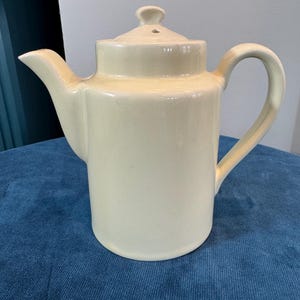 May include: Cream-colored ceramic teapot with a lid and handle. The teapot has a spout for pouring and a rounded body. The handle is curved, and the lid has a small knob. The teapot is sitting on a blue surface.