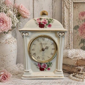 May include: Cream-colored ceramic mantel clock with a gold-tone bezel and Roman numerals. The clock features floral accents in pink and red, with a gold finial. The clock face reads "Formalities".