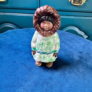 May include: A ceramic figurine of a person wearing a parka with a brown fur-trimmed hood. The parka is white with a green floral pattern and a blue and green geometric band at the waist. The figure has dark hair and wears brown boots and mittens.