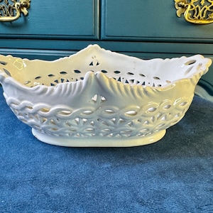 May include: A white ceramic bowl featuring an intricate openwork lattice design along the sides and a scalloped rim. The oval-shaped vessel has a smooth, glossy finish and sits on a solid base, showcasing a detailed woven pattern throughout the body.