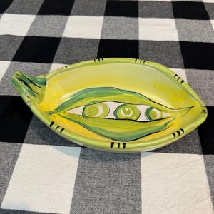 May include: A whimsical, hand-painted ceramic dish in the shape of a pea pod. The dish is primarily yellow with green accents, featuring a stylized design of peas with white and green details. Black lines outline the pod's edges.