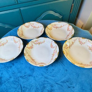 May include: Five white porcelain dishes with a floral design in pink and gold. The dishes are oval-shaped with a scalloped edge. The floral pattern is arranged around the rim of each dish. The dishes are arranged on a blue surface.