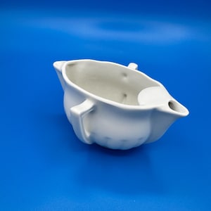 May include: A white ceramic gravy boat featuring a smooth, rounded body with a scalloped base, a single pouring spout, and two small handles on opposite sides. The interior is hollow and the exterior surface is plain, matte, and unadorned.