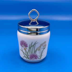 May include: A white ceramic jar with a polished silver-tone metal lid featuring a diamond-shaped handle. The body of the jar is decorated with a botanical illustration of purple chive blossoms and green stems alongside delicate beige wildflower sprigs.