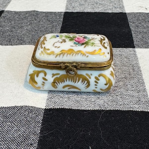 May include: A small, rectangular trinket box with a hinged lid. The box is white with gold accents and a floral design on the lid. The clasp is gold-colored. The box is likely made of porcelain or ceramic.