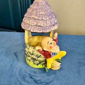 May include: A ceramic figurine of a well with a purple roof and a smiling gnome. The gnome is wearing a red jacket, yellow pants, and a blue shirt. The well has a stone base and a small wooden bucket.
