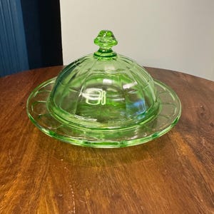 May include: A green glass butter dish with a domed lid and a round base. The lid has a decorative knob on top and a grid-like pattern. The dish is made of clear, light green glass and sits on a wooden surface.