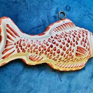 May include: A ceramic fish-shaped mold featuring a glossy white finish with hand-painted reddish-brown details highlighting the scales, fins, and tail. A small metal ring is attached to the top edge for hanging, providing a decorative wall accent.