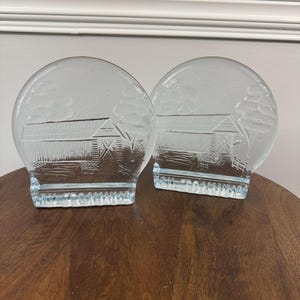 May include: Two clear glass bookends with a barn scene etched on the front. The bookends have a rounded top and a rectangular base. The barn scene includes a building, trees, and a sky. The bookends are sitting on a wooden table.