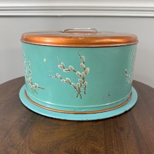 May include: Vintage teal metal cake tin with a copper-colored lid and handle. The tin is decorated with hand-painted white and brown floral sprigs. The tin sits on a matching teal base, perfect for storing baked goods.