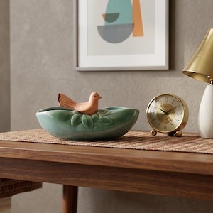 May include: A wooden table displays a teal ceramic bowl with a bird figurine, a gold-toned alarm clock, a white and gold lamp, and two books. A woven runner and artwork are also visible.