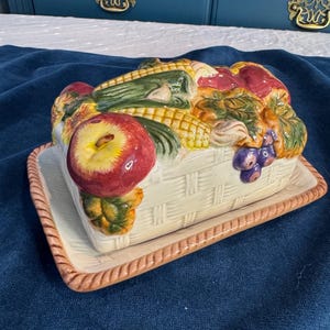 May include: A ceramic butter dish with a basket weave design, topped with a colorful arrangement of fruits and vegetables. The lid features apples, corn, and grapes. The dish sits on a rectangular tray with a brown rope-like border.