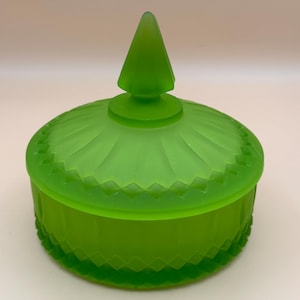 May include: A round, frosted lime green glass jar with a matching lid. The lid features a pointed finial and a radial pattern, while the base and lid edge are decorated with a repeating diamond-shaped geometric texture around the circumference.