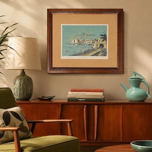 May include: A framed landscape print depicting a coastal scene with a stone archway, boats, and a palm tree, displayed in a dark wood frame with a tan mat. The artwork features a light blue sky and sea, with signed text at the bottom edge.