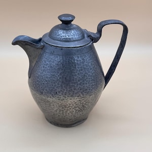 May include: A dark gray metal teapot featuring a hammered texture across its rounded body. The teapot includes a matching lid with a small knob handle, a curved spout, and a sturdy, arched handle attached to the side of the vessel.