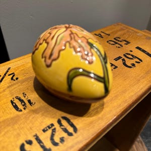 May include: A yellow ceramic egg-shaped decorative object with a brown and green floral design. The object is sitting on a wooden surface with black stenciled numbers and letters.