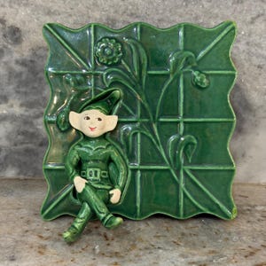 May include: A green ceramic wall hanging featuring a seated elf figure. The elf is wearing a green outfit with a matching hat and has a smiling face. The background is a textured green square with floral and geometric designs.