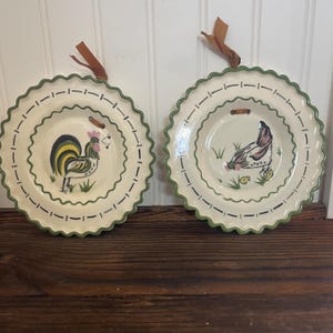 May include: Two decorative ceramic plates with scalloped edges and green trim. One plate features a rooster, the other a hen, both with musical notes. Each plate has a brown leather loop for hanging.