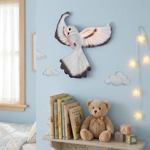 May include: A white and grey wall-mounted bird sculpture with outstretched wings, positioned above a wooden shelf holding a tan teddy bear, several books, and two small alphabet blocks, with painted clouds on the light blue wall.