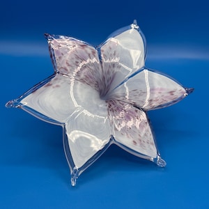 May include: A hand-blown glass sculpture shaped like a five-petaled flower. The glass is primarily clear and white with delicate, scattered purple speckles and swirls concentrated toward the center of the petals, creating a translucent effect.