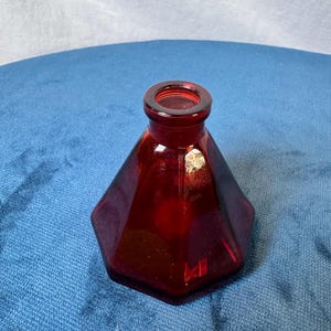 May include: A deep red glass inkwell featuring a faceted hexagonal base that tapers upward to a circular neck and opening. The translucent glass displays a rich crimson hue with smooth, flat sides and a thick, rounded rim at the top.