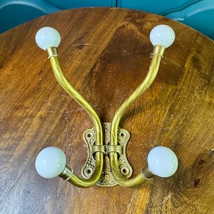 May include: A wall-mounted coat hook featuring a brass-toned metal base with a textured finish and two curved arms. Each arm splits into two ends, totaling four hooks, each capped with a smooth, round, white ball finial for hanging garments.