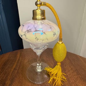 May include: An antique perfume bottle with a bulbous, iridescent glass body and a gold-colored metal top. It features a long, yellow cord with a tassel and a clear glass base. The bottle is displayed on a dark wooden surface.