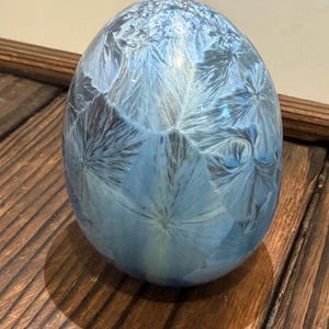 May include: A smooth, egg-shaped ceramic object featuring a crystalline glaze in shades of light and dark blue. The surface displays intricate, star-like patterns radiating from multiple centers, creating a unique visual texture across the entire form.