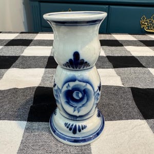 May include: A white ceramic candle holder with blue floral designs. The candle holder has a flared top, a bulbous middle section, and a round base. The blue floral pattern includes a rose design. The candle holder is on a black and white checkered surface.