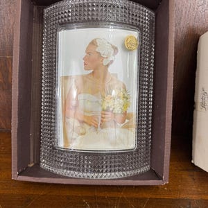 May include: A clear, rectangular photo frame with a textured, diamond-patterned border. The frame holds a photograph of a bride in a white dress, holding a bouquet. The frame is in a brown box, displayed on a wooden surface.