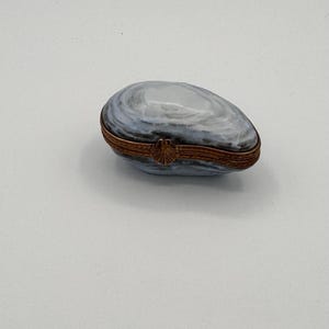 May include: A small, oval-shaped trinket box crafted from polished stone with swirling grey and white patterns. The box features a hinged closure with a metallic, copper-toned band and a small, decorative shell-shaped clasp at the center front.