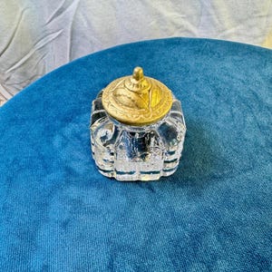 May include: A clear glass jar with a faceted, multi-sided body and a removable, circular brass-toned metal lid. The lid features a textured, embossed pattern with a small, pointed finial at the center, while the glass base has a thick, heavy bottom.
