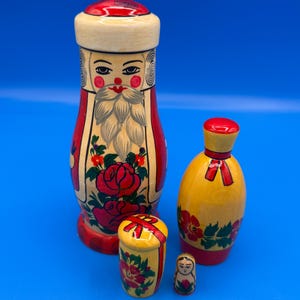 May include: A set of four wooden nesting dolls with hand-painted details. The largest doll features a bearded figure in a red and cream coat with red roses. The smaller dolls are yellow and cream with red floral patterns and red ribbon accents.