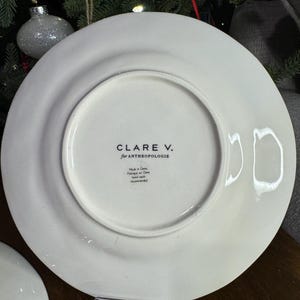 以下が含まれることがあります： 黒で「CLARE V. for ANTHROPOLOGIE」と印刷された白いセラミックプレート。プレートの裏側には、中国製で手洗いを推奨することを示す追加のテキストがあります。プレートは丸く、縁がわずかに盛り上がっています。