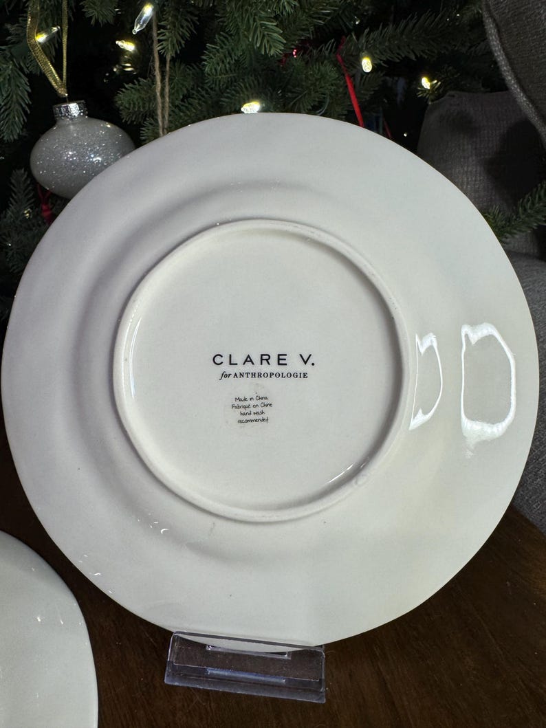 以下が含まれることがあります： 黒で「CLARE V. for ANTHROPOLOGIE」と印刷された白いセラミックプレート。プレートの裏側には、中国製で手洗いを推奨することを示す追加のテキストがあります。プレートは丸く、縁がわずかに盛り上がっています。