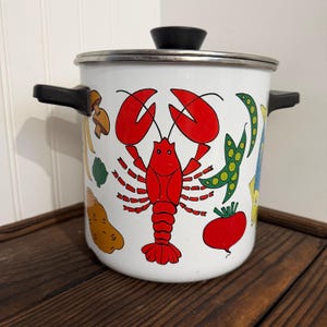 May include: White enamel cooking pot with a black lid and handles. The pot is decorated with a red lobster and colorful vegetables, including peas, a potato, and a beet. The pot has a silver rim.