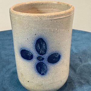 May include: A stoneware tumbler with a light gray speckled finish and a blue floral design. The handmade cup has a rounded base and a slightly flared rim. The blue design is a four-petal flower.